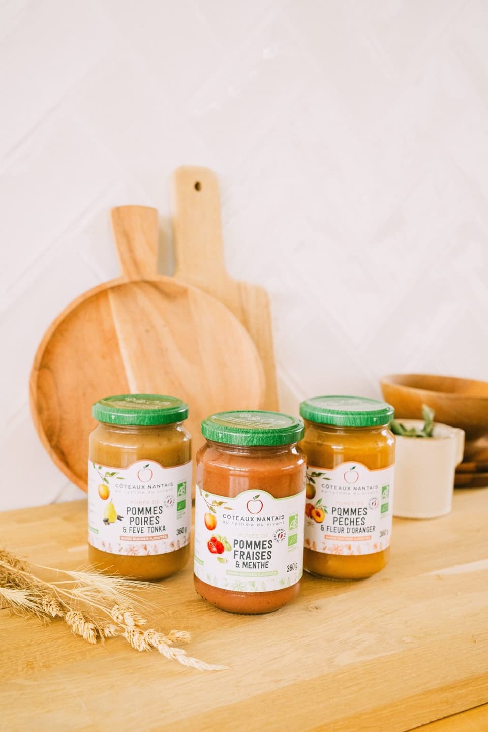 Trois pots de compote, pommes-poires, pommes-fraises et pommes-pêches, posés sur une planche en bois