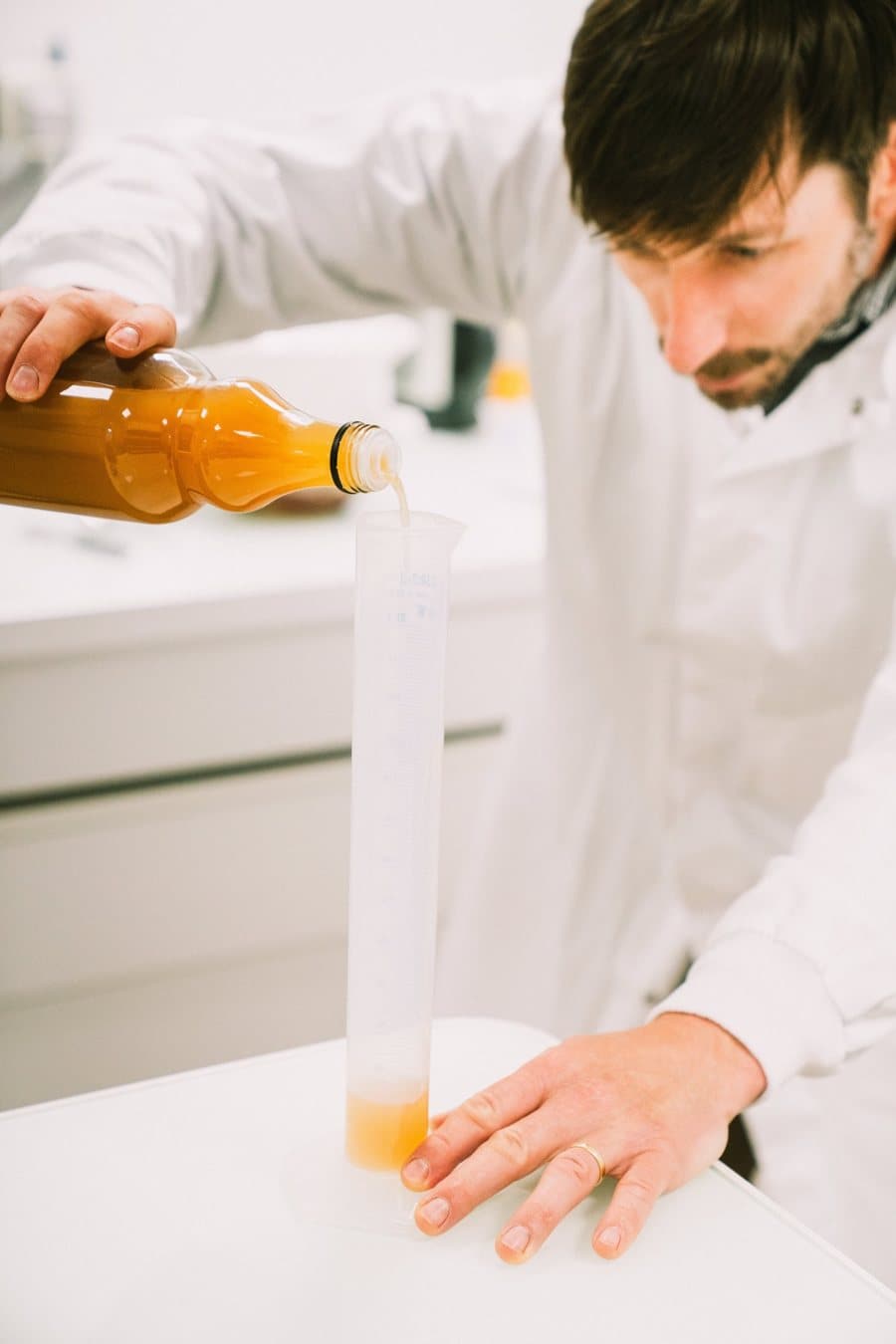 Un technicien en blouse blanche verse du jus de pomme dans une éprouvette graduée pour l’analyser